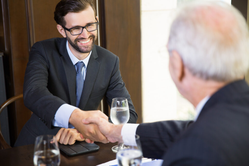 Portrait of young businessman sitting in modern office and shaking hands with his mature partners after finishing meeting