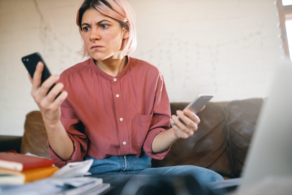 Job burnout. Angry overworked businesswoman in casual clothes trying to cope with freelance work because of social distancing, holding two cell phones ringing, being buried with multiple tasks