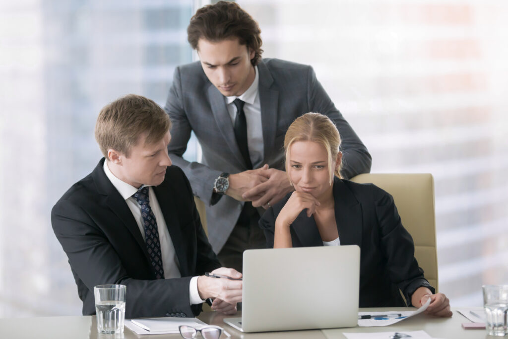 Group of three business partners discussing new project at meeting in office room, using laptop. Young confident businessman in formal wear suit explaining idea and showing presentation on screen